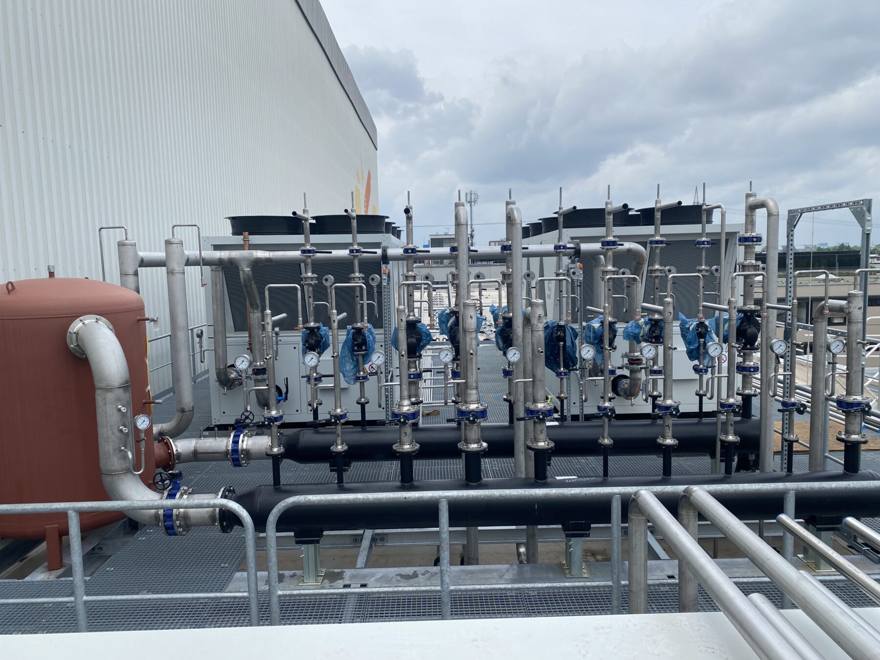 Detailansicht einer technischen Anlage auf einem Dach, bestehend aus einem komplexen System von Rohren, Pumpen, Manometern und einem großen braunen Tank unter einem bewölkten Himmel.