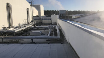 Dach einer Industrieanlage mit Ammoniak-Rohrleitungen und einem dampfenden Rückkühler an einem kalten, sonnigen Wintertag.